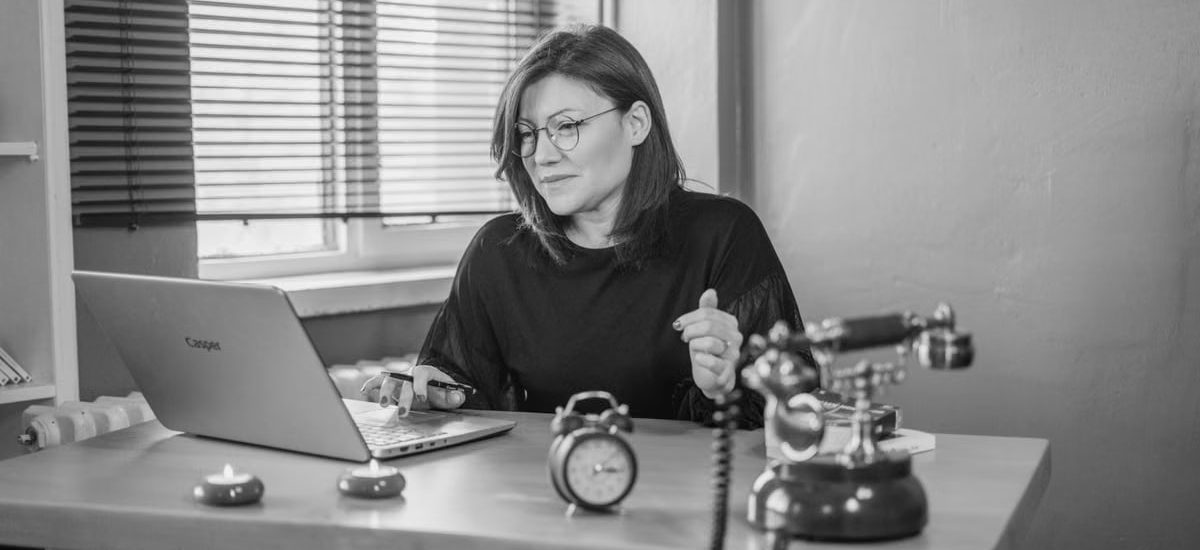 Black and white photo of a woman working at a desk with a laptop and vintage telephone.
