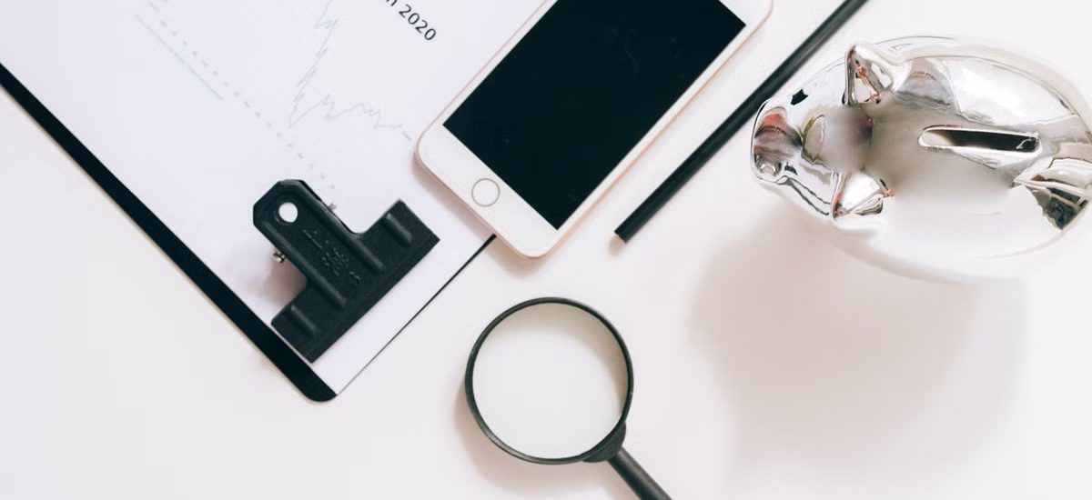 A flat lay of financial documents with a smartphone, magnifying glass, and piggy bank.